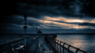 This architectural image captures a long wooden pier extending into the ocean under a dramatic, clou