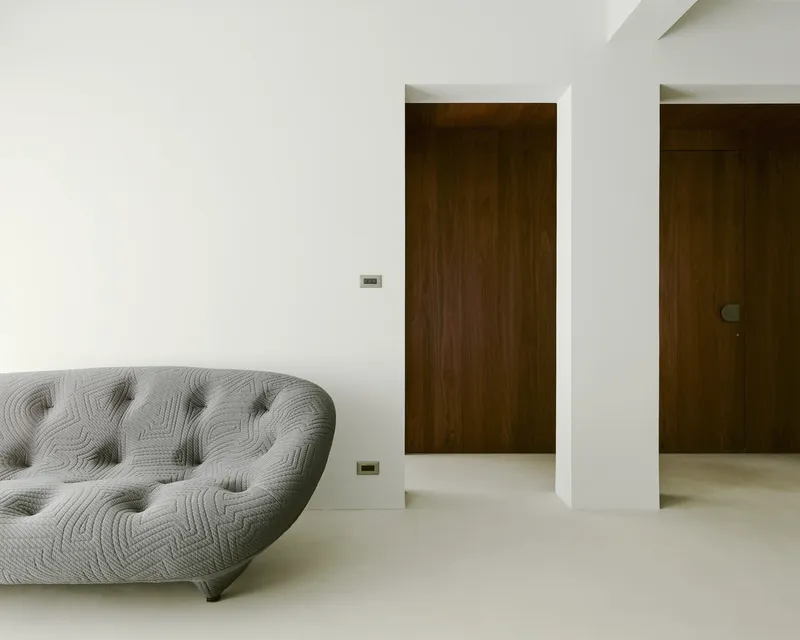 Tufted grey sofa against white wall with two walnut doorways receding into the distance