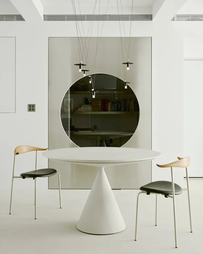 Dining table with conical white base and two chairs beneath pendant lights and a circular mirror