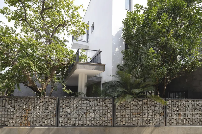 White facade with cantilevered balconies above a gabion retaining wall and mature trees