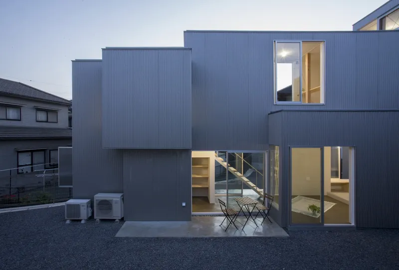 Rear facade with corrugated metal cladding and glazed openings overlooking a gravel courtyard at dusk