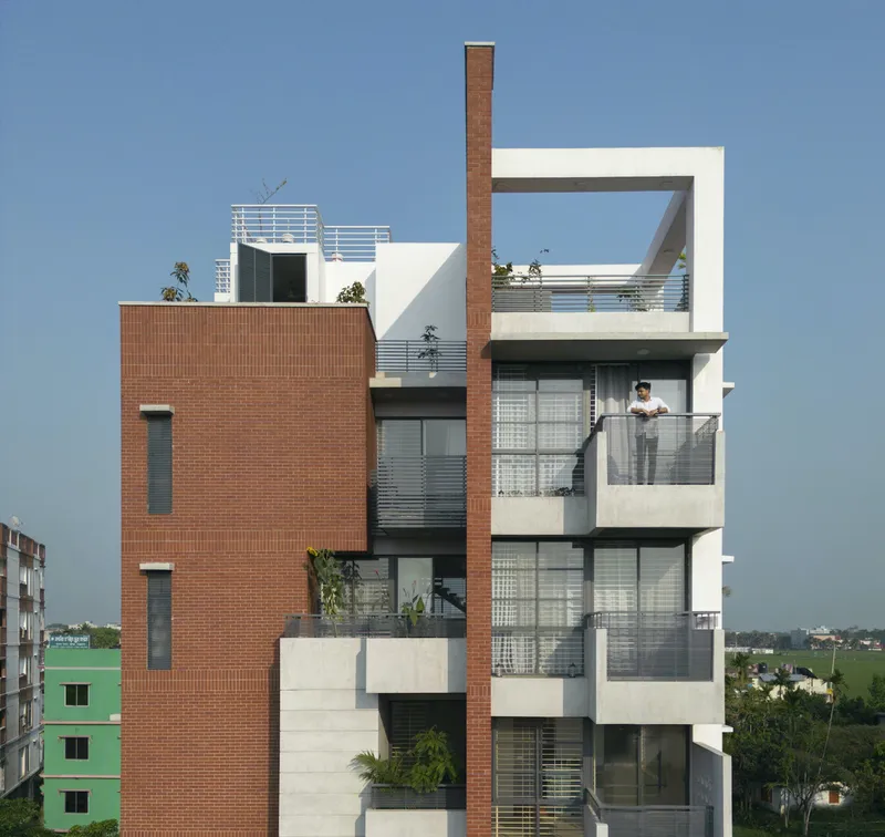 Street facade combining red brick volumes and white concrete balconies with metal railings under clear blue sky