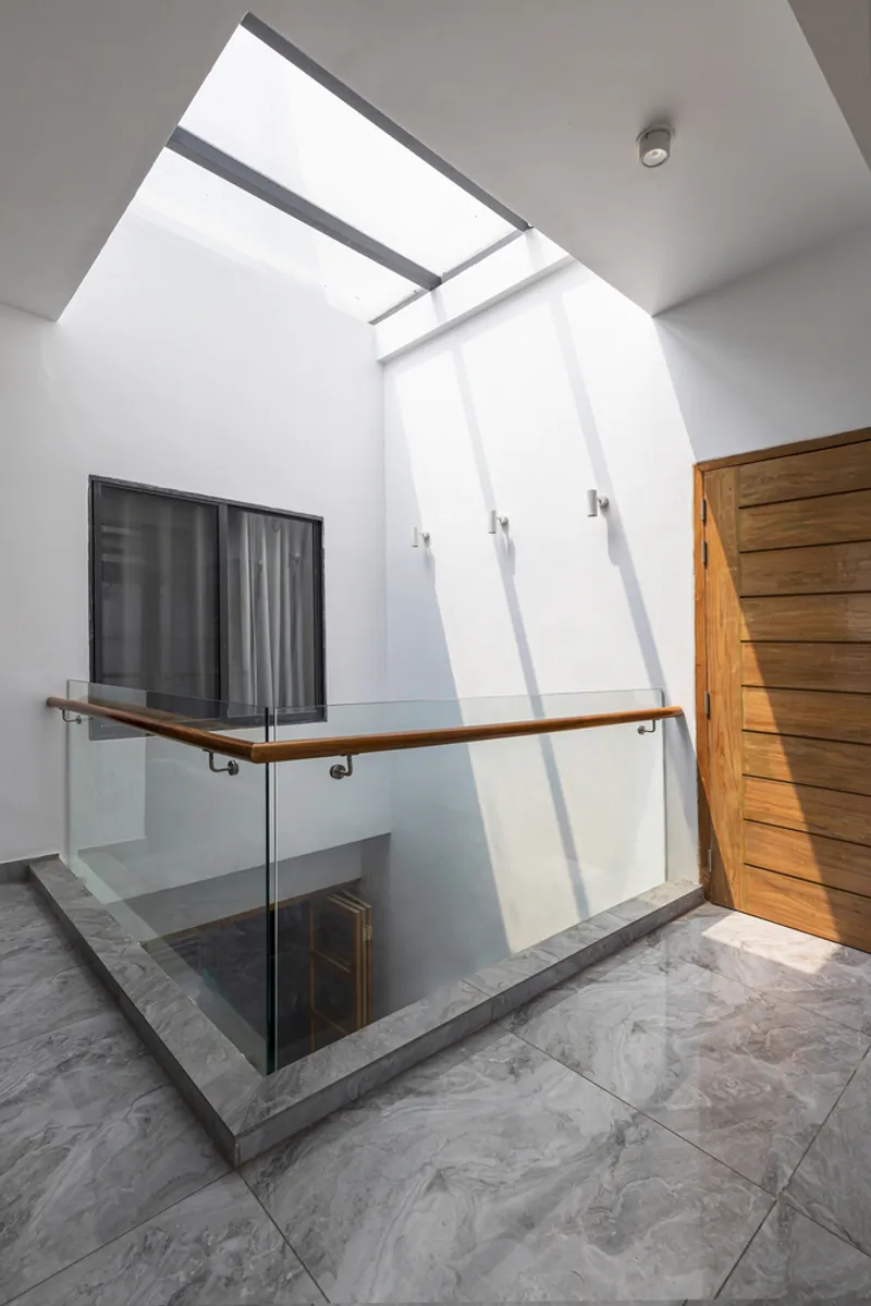 Upper landing with glass balustrade and timber handrail beneath a skylight casting geometric shadows across white walls