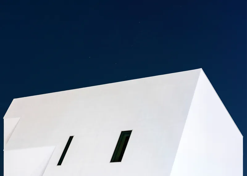 Upward view of white facade with two angled window openings under a deep blue evening sky