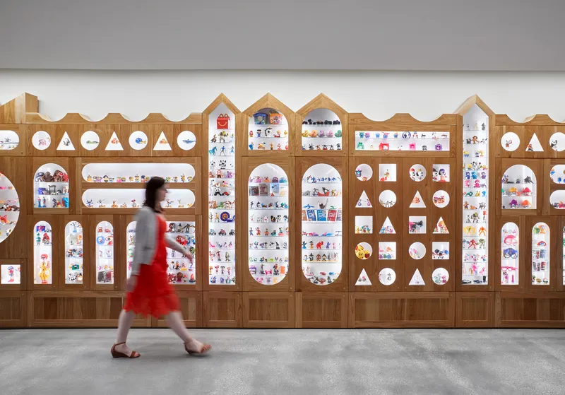 Timber display wall with arched and circular cutouts showcasing colorful objects as a person in red walks past