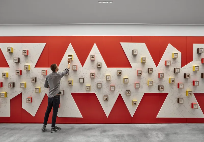 Red and white zigzag wall installation displaying small framed objects as a person reaches toward it