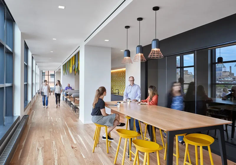 Open office corridor with timber communal table and yellow stools alongside glazed meeting rooms