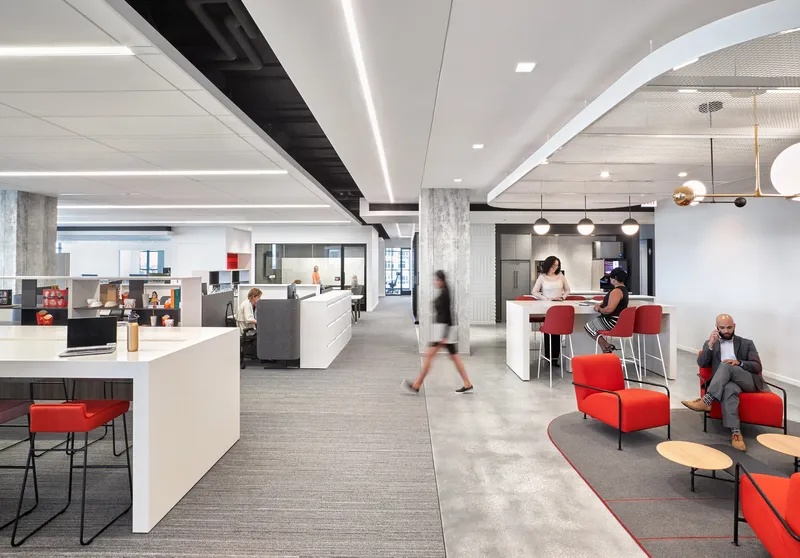 Open office floor with linear ceiling lighting and people moving between workstations and lounge seating