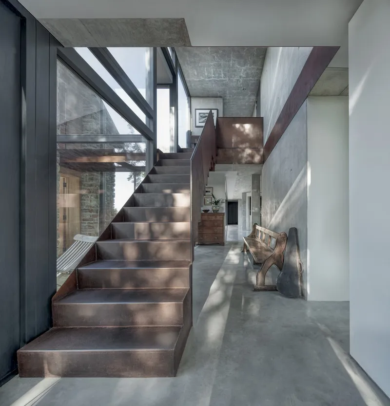 Steel staircase with copper-toned treads ascending through a double-height space with exposed concrete and natural light