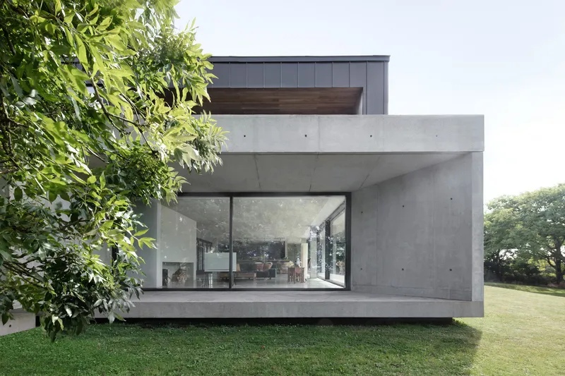 Corner view of the concrete volume with large glass openings framed by green lawn and trees