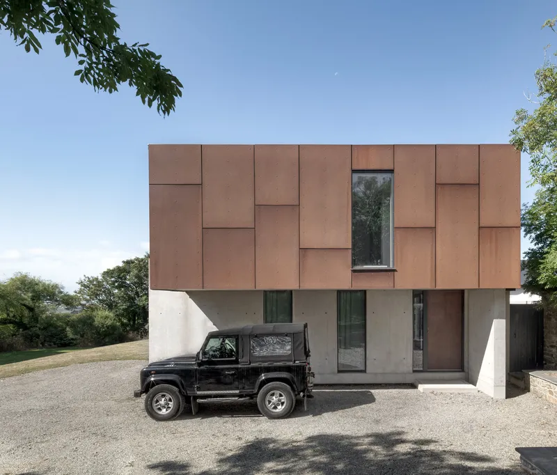Street facade with offset copper panels above concrete base and parked vehicle in gravel driveway
