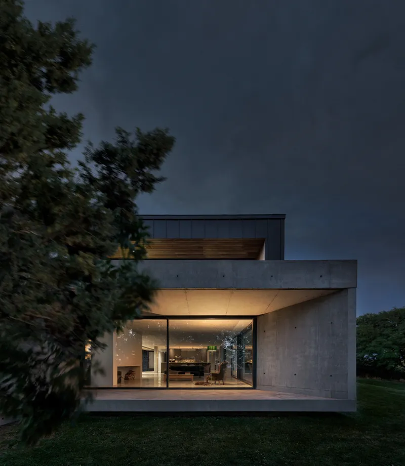 Cantilevered concrete terrace and volume with full-height glazing opening to the living space at dusk