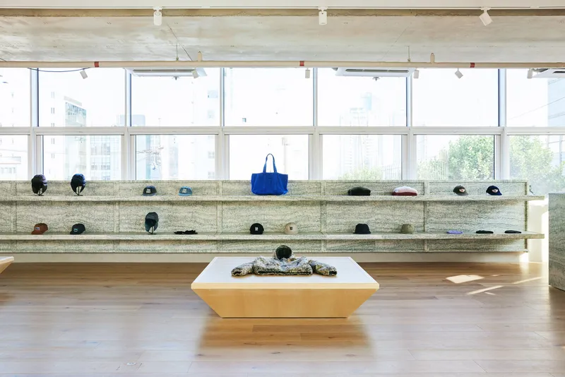 Accessories wall: long granite shelf with integrated niches displaying caps and bags, plywood table with folded garments, blue tote accent