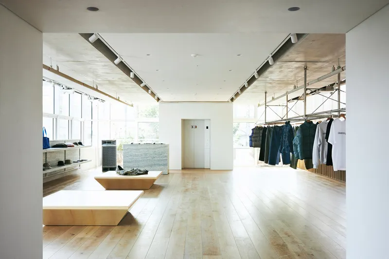 Full store view: denim and outerwear on hanging rails, plywood tables, granite counter, white walls, concrete ceiling, wide oak floor