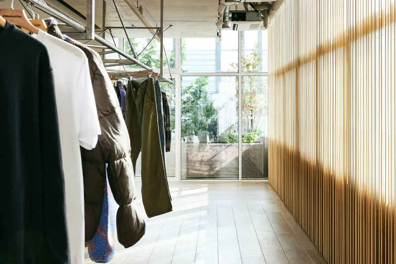 Window and hanging rail: garments along the glass, tree and terrace visible outside, timber slat wall catching light
