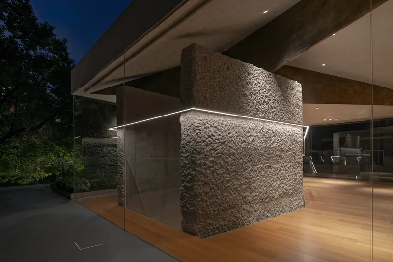 Night detail: rough stone stair core visible through glass, LED strip glowing, oak floor, concrete beam, garden behind