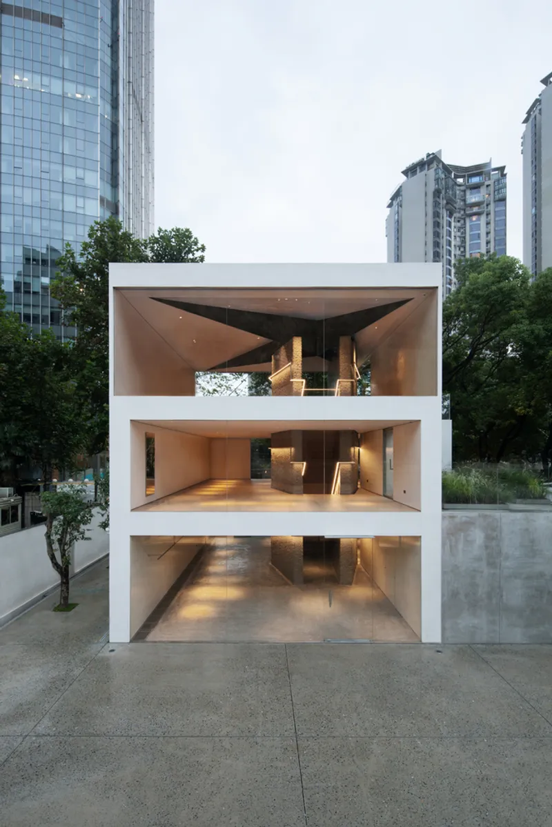 Exterior at dusk: white two-storey cube on pilotis, oak floors and gallery visible through frameless glass, Shanghai skyline behind