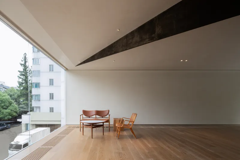 Upper gallery: oak floor, white walls, dark angular ceiling beam, two leather chairs and coffee table, frameless glass wall to city view