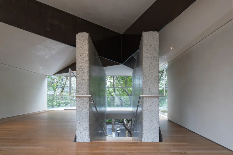 Central staircase: rough granite columns, glass balustrade, white treads, double-height space, mature trees visible through glass