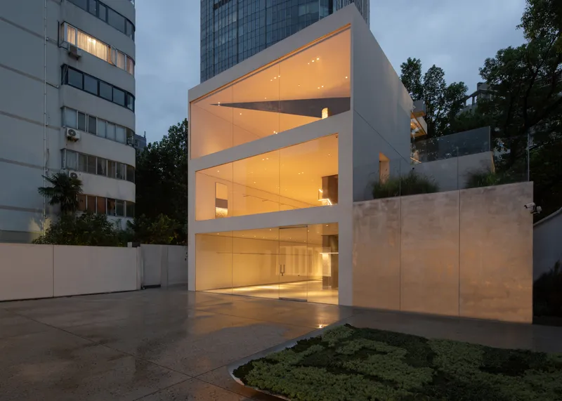 Exterior at dusk: white cube glowing from within, diagonal floor slabs visible through glazing, concrete forecourt, city buildings behind