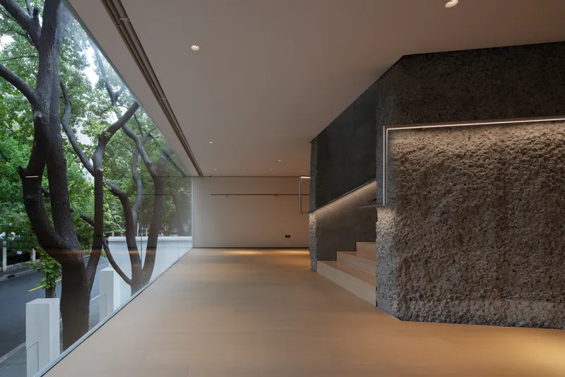 Ground floor gallery: rough stone core, LED handrail line, frameless glass with mature plane trees outside, oak floor