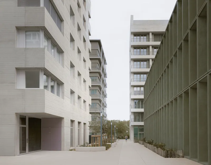 Pedestrian pathway between concrete residential facades and a green metal screen wall with planted beds