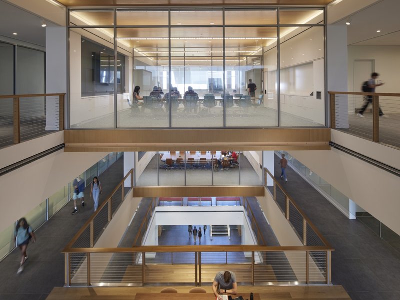 Multi-story atrium with glass-walled conference rooms and timber-clad bridges connecting the upper levels