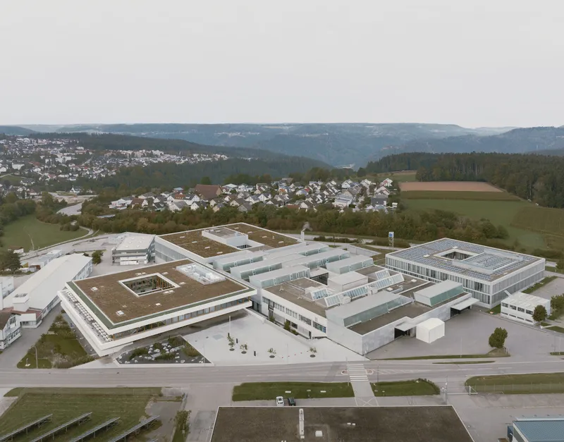 Aerial view of the clustered white volumes with green roofs across the sloped landscape