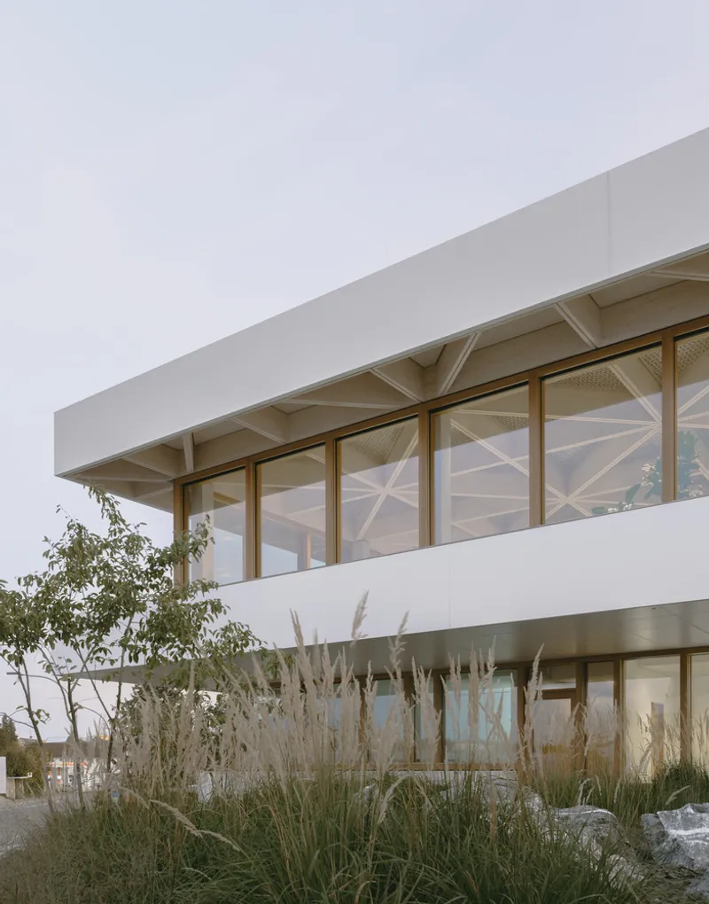 Two-story glazed facade with timber frames and exposed ceiling structure behind planted beds of tall grasses