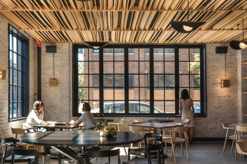 Dining room: timber slat ceiling, exposed cream brick wall, large black steel windows, oak tables and chairs, pendant fan lights