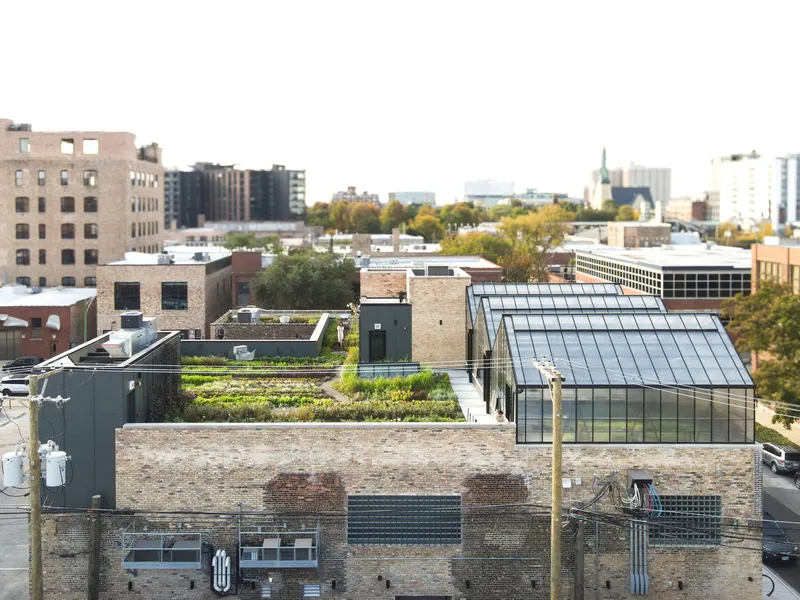 Elevated view: rooftop farm and greenhouses from above, green planting beds, city skyline with autumn foliage