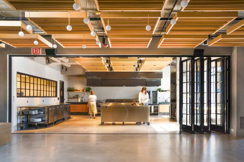 Test kitchen: timber beam ceiling, stainless steel island, figure working, black bifold doors to dining area, globe lights