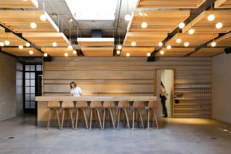 Tasting bar: long timber counter with stools, horizontal timber slat wall, wine storage at right, skylight above, globe lights
