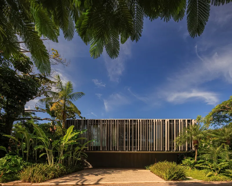 Approach: vertical timber brise-soleil facade behind tropical planting, stone path, blue sky above