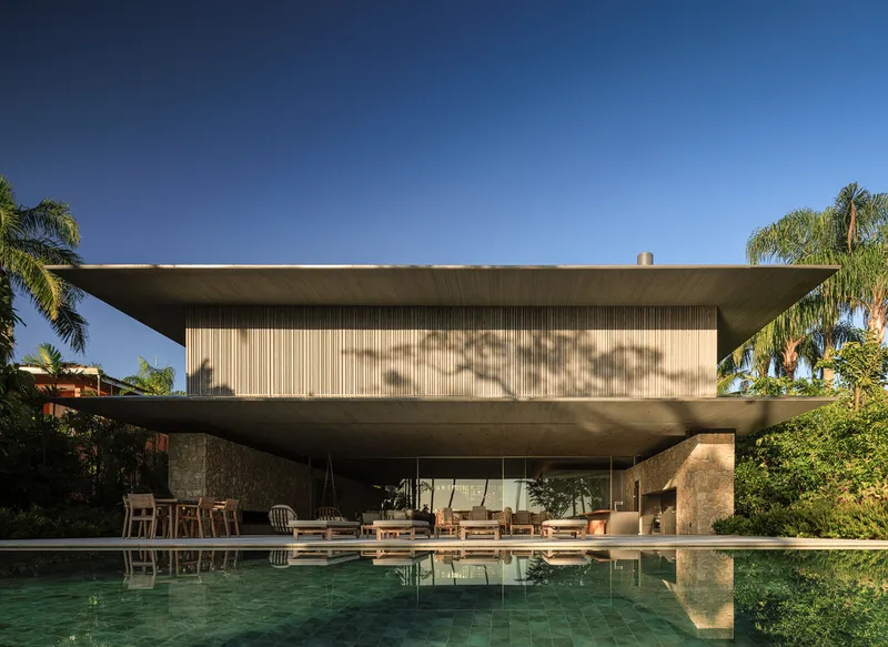 Pool view: two-storey house with deep cantilevered concrete roof, timber slats above stone wall, pool reflecting the facade, palm trees