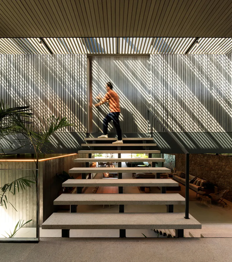 Interior staircase: floating concrete treads, timber brise-soleil screen behind casting striped shadows, figure ascending