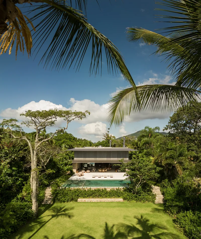 From the garden: house as a horizontal bar above the pool, lawn in foreground, palm trees and tropical canopy framing