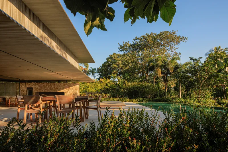 Pool terrace at golden hour: deep concrete roof overhang, timber dining table and chairs, curved pool edge, tropical planting