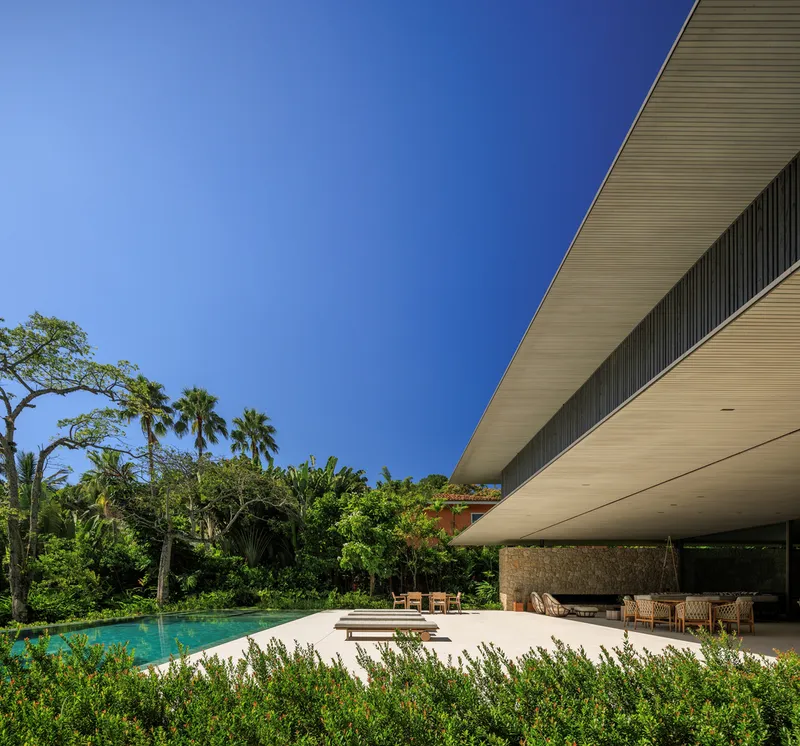 Side view: deep concrete cantilever over stone deck, pool, sun loungers, tropical planting, clear blue sky