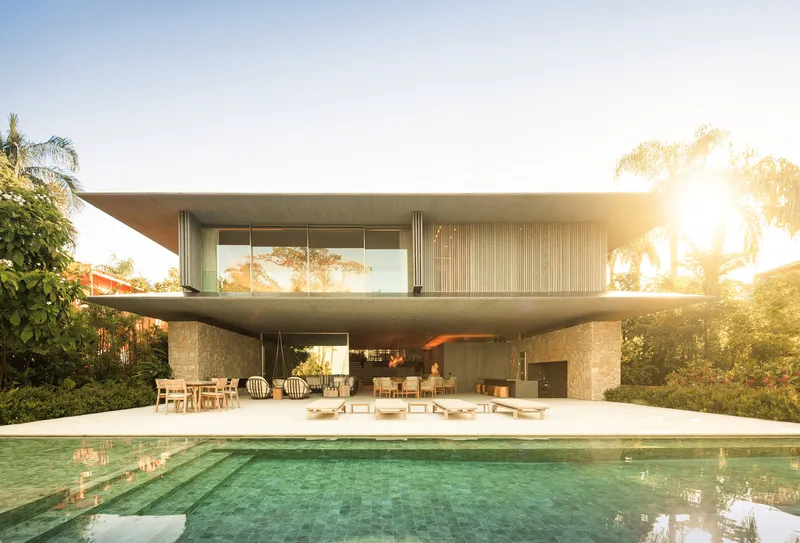 Pool facade at sunset: stone wall ground floor, timber-slatted upper floor, pool, sun flare through the trees