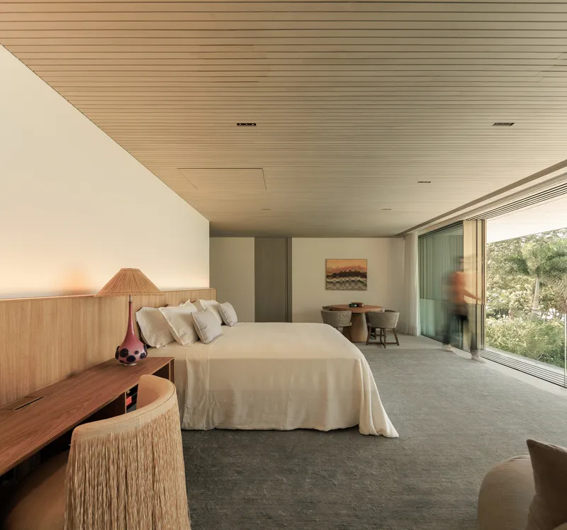 Bedroom: timber ceiling, white bedding, timber headboard wall, rattan armchair, artwork, floor-to-ceiling glass to garden