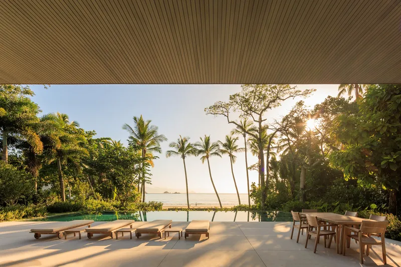 Pool terrace at sunset: timber ceiling soffit, sun loungers, dining table, palm trees, ocean and beach visible, golden light