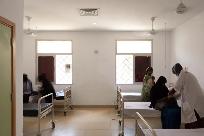 Ward interior: hospital beds, patients and medical staff, ceiling fans, square windows with timber shutters, white walls