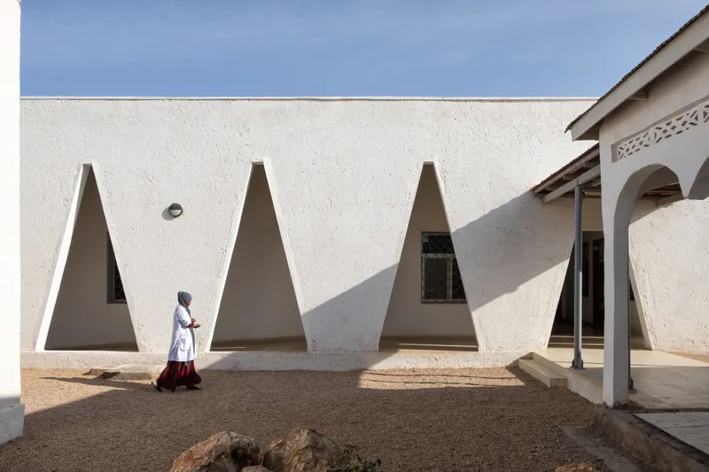 Courtyard wall: row of triangular openings, figure in white sitting at the base, older hospital building visible at right