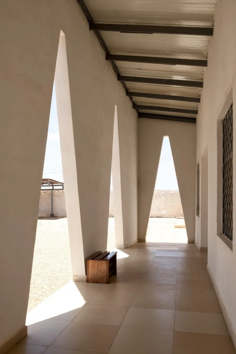 Covered corridor: row of triangular openings casting sharp light on the floor, timber roof beams and rafters above