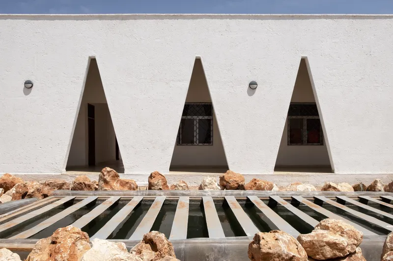 Facade detail: three triangular windows above a slatted water channel with local rocks, wall-mounted light