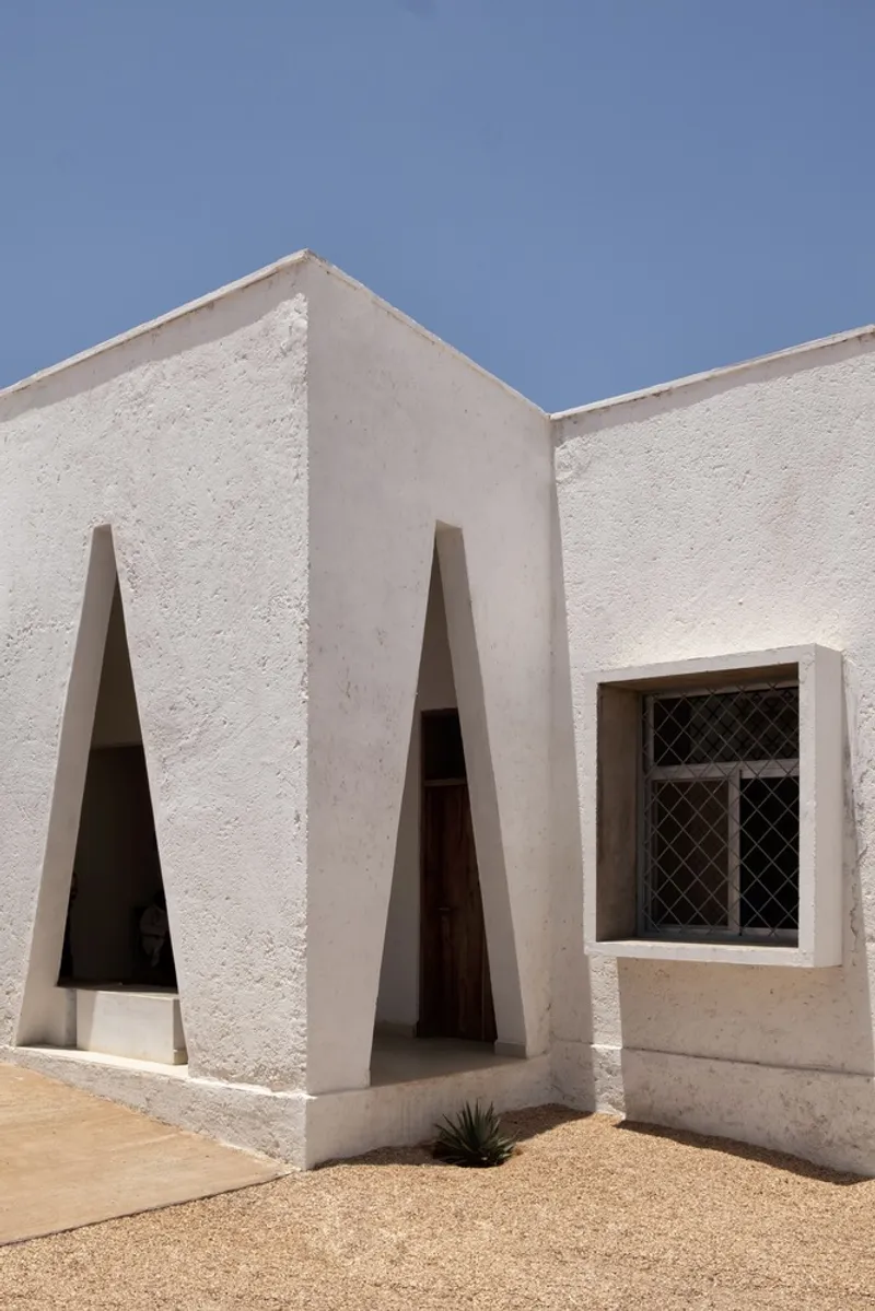 Building corner: two white volumes meeting at right angles, triangular windows on both faces, desert ground