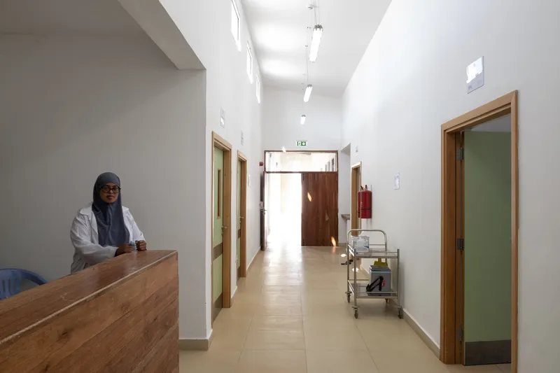 Interior corridor: nurse at timber reception desk, timber doors to wards, clerestory light strip along the ceiling