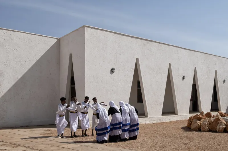 Community gathering: women in white traditional dress assembled beside the wall with triangular openings