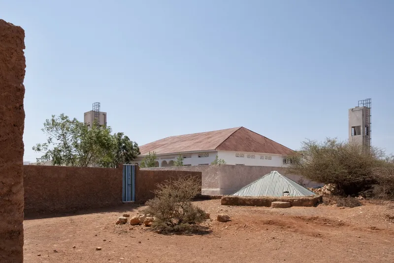 Existing hospital: older building with corrugated metal roof and water towers, dry desert terrain around